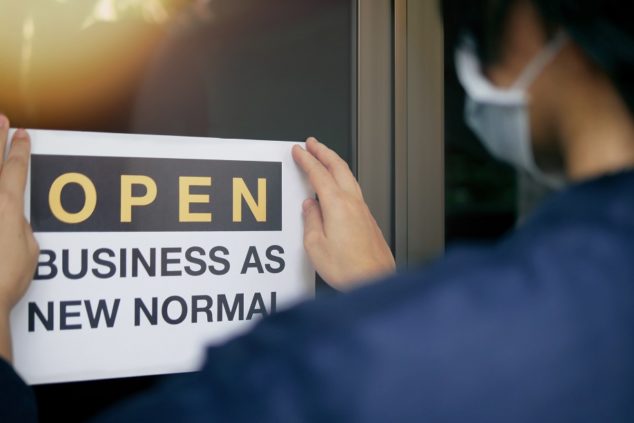 Reopening for business adapt to new normal in the novel Coronavirus COVID-19 pandemic. Rear view of business owner wearing medical mask placing open sign “OPEN BUSINESS AS NEW NORMAL” on front door.